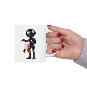 May include: White ceramic mug featuring a glossy black alien figure holding a small dog. The alien has large, expressive eyes. The dog is brown and white. The mug has a white handle and is held by a person with red nail polish.