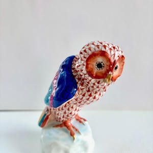 May include: A ceramic owl figurine with a red, white, and blue color scheme. The owl has a detailed pattern on its head and body, with a blue wing and a white base. The owl's eyes are black, and its beak and talons are gold.