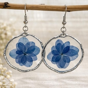 May include: A pair of circular earrings with pressed blue flowers set in clear resin. The earrings have a silver-coloured metal frame and hook fastenings, displaying a delicate floral design. The earrings are presented against a neutral backdrop.