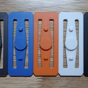 May include: A collection of five 3D-printed fidget spinners in a row. The spinners are in black, blue, orange, white, and black. Each spinner has a central disc and a rectangular frame with cutouts.