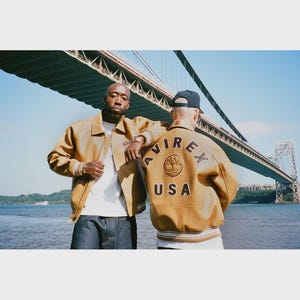 May include: Two people wearing tan leather jackets with the word "AVIREX" and "USA" on the back. The jackets have a classic bomber style. They are standing near a body of water with a bridge in the background.