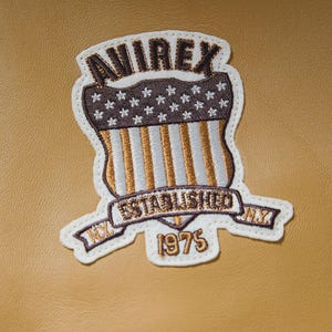 May include: Embroidered patch with the text "AVIREX" at the top, a shield design with stars and stripes, and the words "ESTABLISHED NY 1975". The patch has a white border and is set against a tan background.