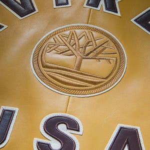 May include: Close-up of a mustard yellow leather jacket with brown and white embroidered patches. The jacket features a circular patch with a tree design and the letter patches spelling out "USA".