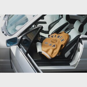 May include: A tan leather jacket with the text "AVIREX USA" is inside a white and black car. The jacket has a cream-colored collar and cuffs. The car's interior features white and black leather seats and a silver exterior.