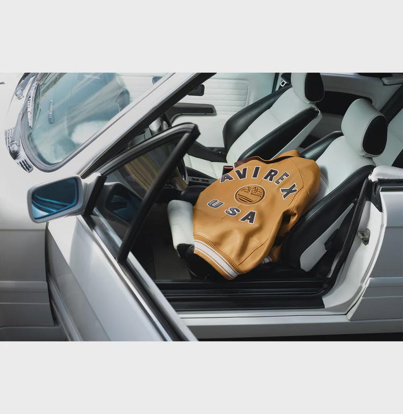 May include: A tan leather jacket with the text "AVIREX USA" is inside a white and black car. The jacket has a cream-colored collar and cuffs. The car's interior features white and black leather seats and a silver exterior.