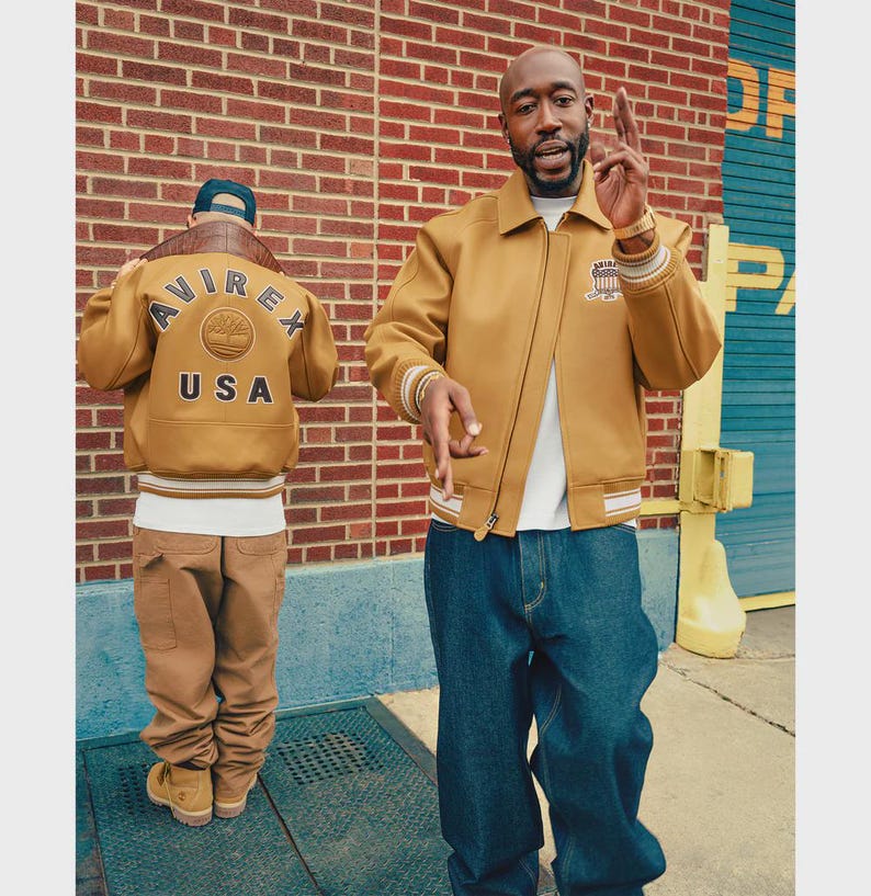 May include: Two people wearing tan bomber jackets and blue jeans stand in front of a brick wall. One jacket has "AVIREX USA" on the back. The other jacket has a patch on the chest. The jackets have white and tan striped cuffs.