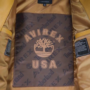 May include: Mustard yellow leather jacket interior with a brown lining. The lining features the Avirex and Timberland logos, along with the text "USA". The jacket has a classic design.
