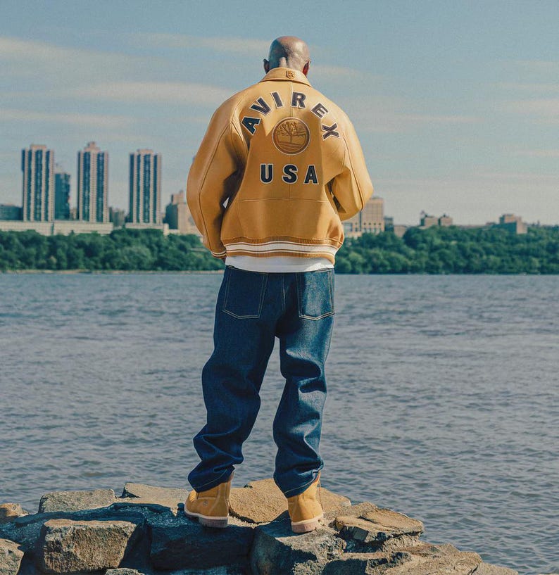 May include: A person wearing a mustard yellow leather jacket with the word "AVIREX" arched across the back, above a circular emblem and the word "USA". The person is also wearing blue jeans and tan boots, standing on rocks near a body of water.