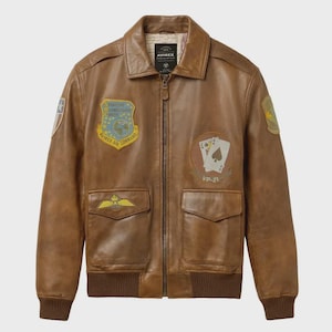 Puede incluir: Chaqueta de cuero marrón con cremallera, dos bolsillos delanteros y puños y dobladillo acanalados. La chaqueta presenta parches con texto y gráficos, incluido el logotipo de Avirex Air Command e imágenes de naipes. Estilo bomber clásico.