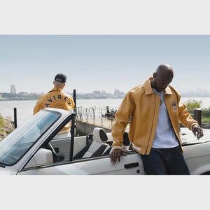 May include: Mustard yellow leather jacket with white trim and a small patch on the chest. The jacket is worn by a person leaning on a white convertible car. Another person is in the background wearing a similar jacket.