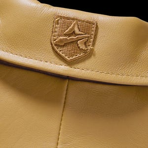 May include: Close-up of a tan leather garment with a stitched emblem. The emblem is a detailed, embroidered patch in shades of tan and brown, featuring a stylized design. The leather has a subtle texture and a warm, neutral tone.