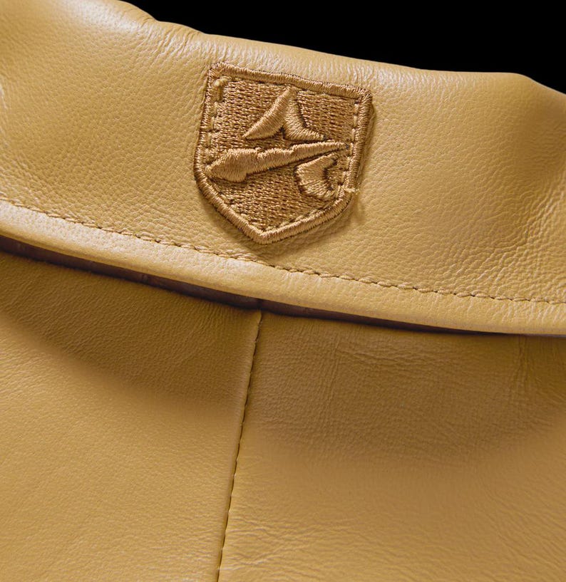 May include: Close-up of a tan leather garment with a stitched emblem. The emblem is a detailed, embroidered patch in shades of tan and brown, featuring a stylized design. The leather has a subtle texture and a warm, neutral tone.
