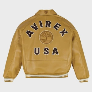Puede incluir: Chaqueta de cuero color canela con la palabra "AVIREX" en letras marrones en la parte superior de la espalda, encima de un emblema circular y la palabra "USA". La chaqueta tiene cuello, puños acanalados y dobladillo acanalado con rayas blancas.