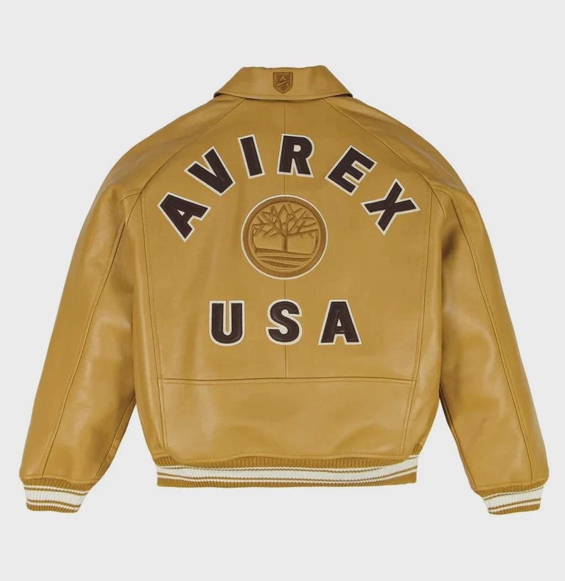 May include: A tan leather jacket with the word "AVIREX" arched across the upper back in brown lettering, above a circular emblem and the word "USA". The jacket has a collared neckline, ribbed cuffs, and a ribbed hemline with white stripes.