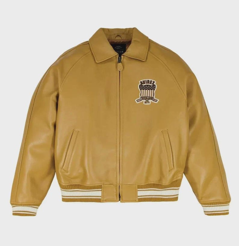May include: Mustard-colored leather jacket with a collared neckline and a zipper closure. Features a patch on the upper left chest with the text "AVIREX" and a design. The jacket has two front pockets and ribbed cuffs and hem with white stripes.