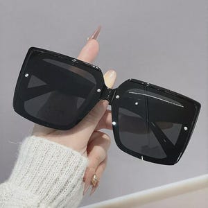May include: Black square sunglasses with dark lenses. The sunglasses have small, silver accents on the corners of the frames. The frames are held by a hand wearing a white sweater.