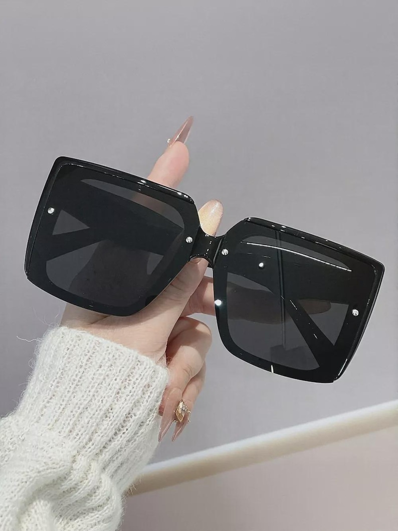May include: Black square sunglasses with dark lenses. The sunglasses have small, silver accents on the corners of the frames. The frames are held by a hand wearing a white sweater.
