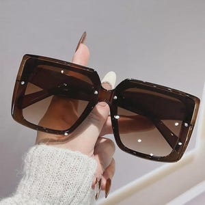 May include: Brown square sunglasses with gradient lenses. The sunglasses have a thick frame and are embellished with small, white studs. The lenses transition from a darker shade at the top to a lighter shade at the bottom.