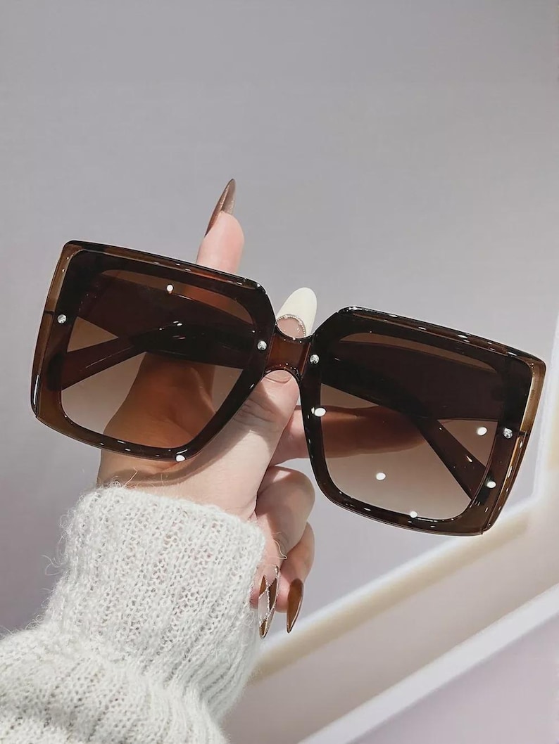 May include: Brown square sunglasses with gradient lenses. The sunglasses have a thick frame and are embellished with small, white studs. The lenses transition from a darker shade at the top to a lighter shade at the bottom.