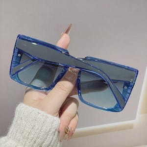 May include: Blue square-framed sunglasses with gradient lenses. The sunglasses have a thick frame and a modern design. The lenses transition from a darker shade at the top to a lighter shade at the bottom. The sunglasses are held by a person.