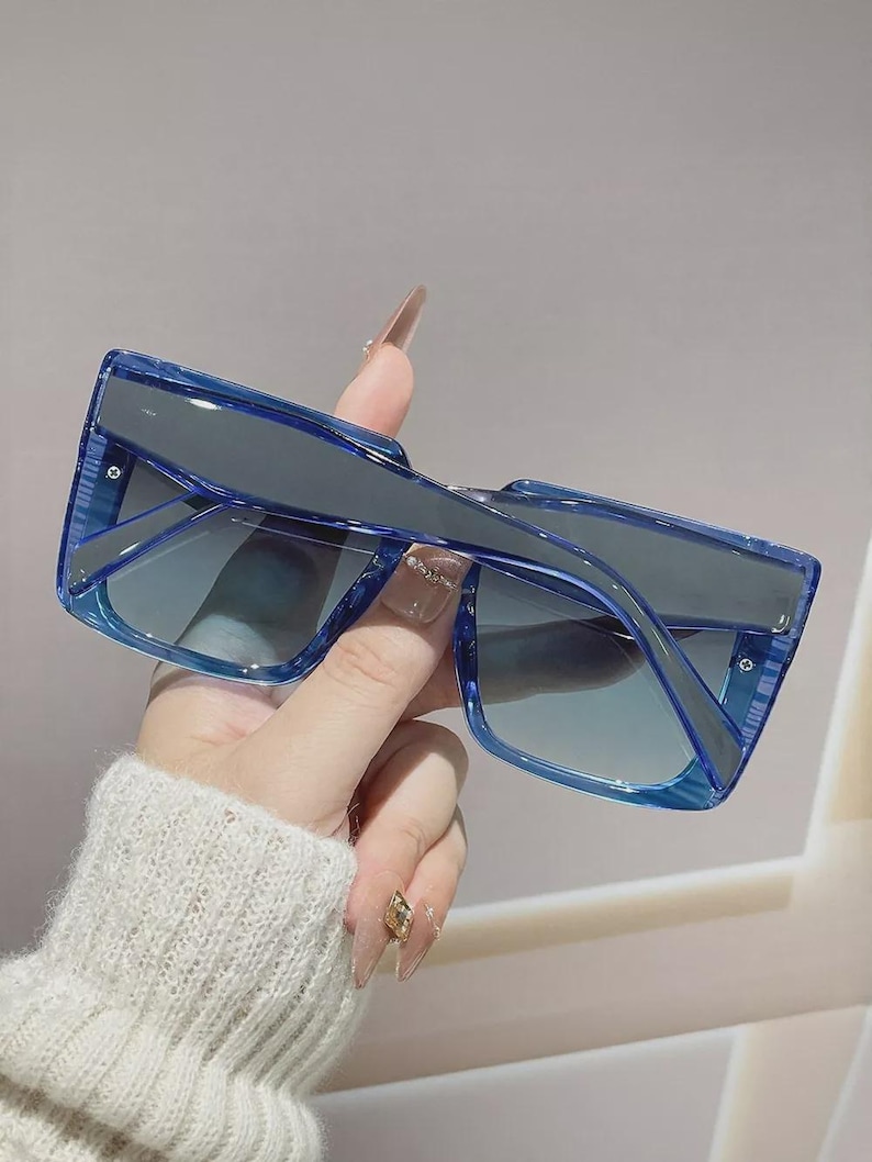 May include: Blue square-framed sunglasses with gradient lenses. The sunglasses have a thick frame and a modern design. The lenses transition from a darker shade at the top to a lighter shade at the bottom. The sunglasses are held by a person.