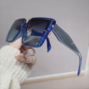 May include: Blue square-framed sunglasses with gradient lenses. The sunglasses have a thick, transparent frame and dark lenses. The arms of the sunglasses are also blue. The sunglasses are held up by a hand.