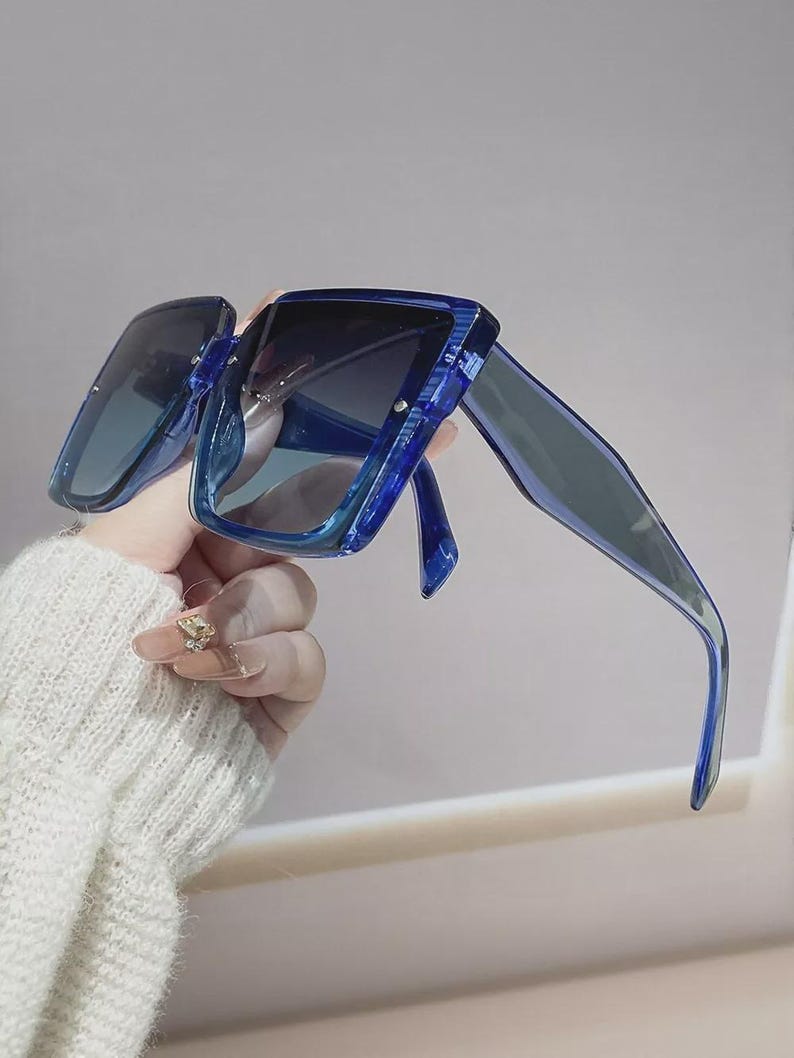 May include: Blue square-framed sunglasses with gradient lenses. The sunglasses have a thick, transparent frame and dark lenses. The arms of the sunglasses are also blue. The sunglasses are held up by a hand.