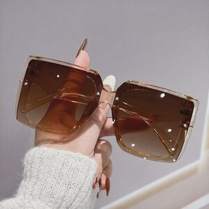 May include: Brown square sunglasses with a gradient tint. The sunglasses have a clear frame and are embellished with small, white, circular accents. The lenses are a darker shade of brown at the top, fading to a lighter shade at the bottom.