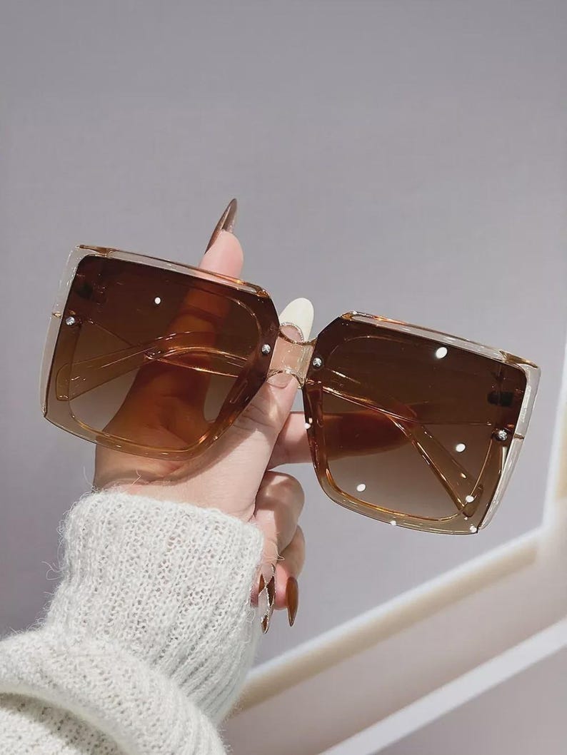 May include: Brown square sunglasses with a gradient tint. The sunglasses have a clear frame and are embellished with small, white, circular accents. The lenses are a darker shade of brown at the top, fading to a lighter shade at the bottom.