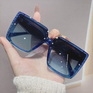 May include: Oversized, square sunglasses in a striking blue colour. The sunglasses have a bold, geometric design with dark lenses. The frames have a textured appearance, and small silver accents are visible on the corners. A fashionable accessory for eye protection.