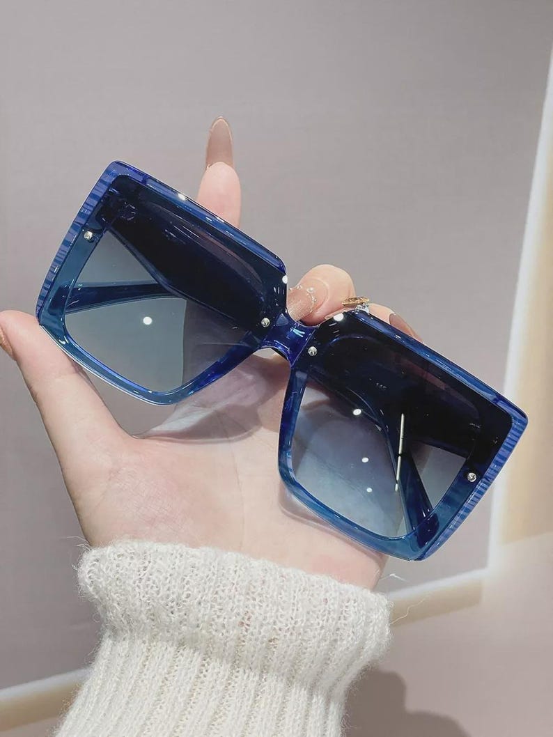 May include: Oversized, square sunglasses in a striking blue colour. The sunglasses have a bold, geometric design with dark lenses. The frames have a textured appearance, and small silver accents are visible on the corners. A fashionable accessory for eye protection.