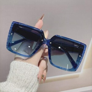 May include: Blue square sunglasses with gradient lenses. The sunglasses have a thick frame and are held up by a hand wearing a white sweater. The lenses transition from dark to light blue.