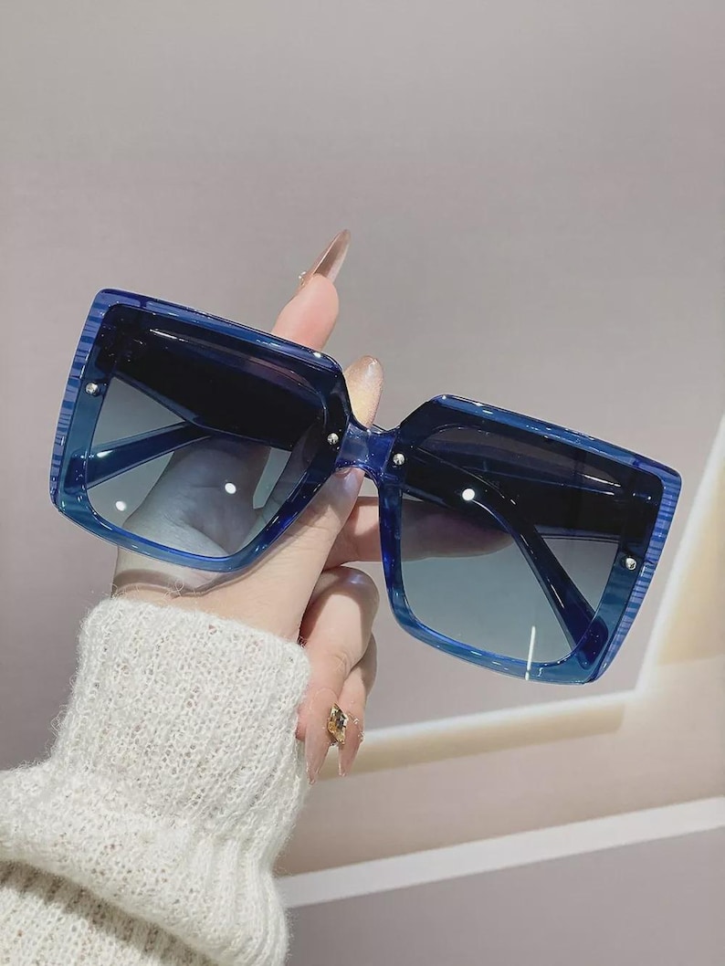 May include: Blue square sunglasses with gradient lenses. The sunglasses have a thick frame and are held up by a hand wearing a white sweater. The lenses transition from dark to light blue.