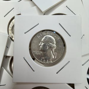May include: A collection of silver quarter coins, each encased in a white cardboard holder. The coins feature the profile of George Washington and the year 1964. The holders are secured with metal staples.