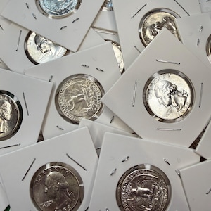 Könnte beinhalten: Eine Sammlung silberner Vierteldollar-Münzen, jeweils in einem weißen Kartonhalter. Die Münzen zeigen Porträts und Adlerdesigns mit sichtbaren Daten. Die Halter sind mit Metallklammern befestigt, was eine ordentliche Präsentation ergibt.