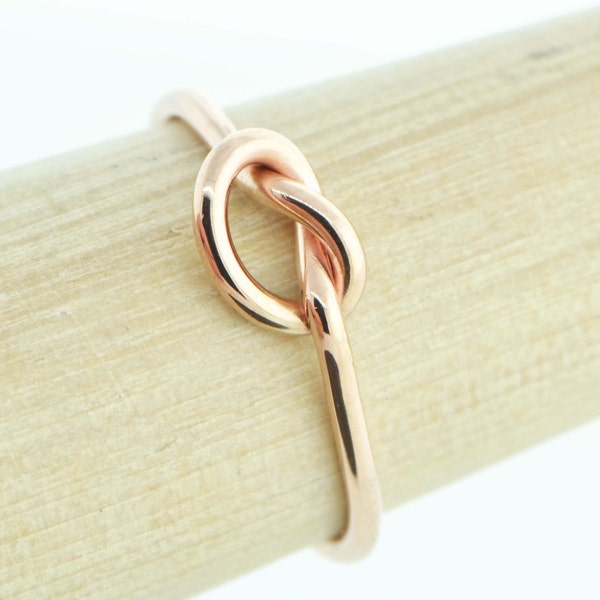 May include: A polished rose gold ring featuring a central knot design. The band is smooth and circular, showcasing a warm metallic finish that reflects light across its surface. The knot is formed by the metal looping over itself in a continuous form.