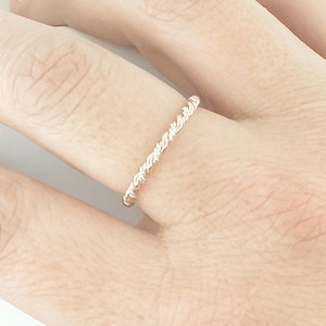 May include: A delicate, twisted silver ring with a subtle rose gold accent. The ring is worn on a finger.