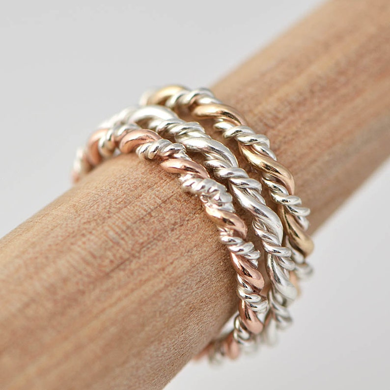 May include: A set of three twisted rings, one silver, one gold, and one rose gold. The rings are stacked on top of each other and are worn on a finger.