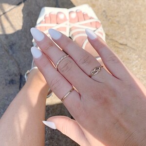 May include: A hand with white nail polish is wearing three gold rings. The rings are a thin band, a twisted band, and a knot design.