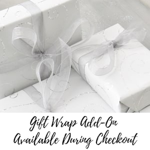 May include: White gift boxes with silver ribbon and a silver swirl pattern. The text "Gift Wrap Add-On Available During Checkout" is displayed below the boxes.