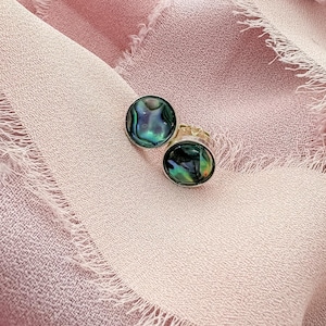 May include: A pair of silver-toned stud earrings with iridescent abalone shell inlays. One earring is round, the other is oval. The abalone shell displays shades of green, blue, and purple. The earrings are set against a pink fabric background.