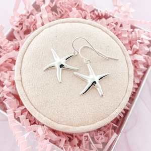 May include: A pair of silver starfish earrings displayed on a beige circular pad inside a silver gift box with pink paper filler. The earrings have a hook design and are made of a shiny, reflective metal.
