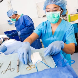 May include: An operating room scene with two medical professionals in blue scrubs, caps, and masks. Surgical instruments, including scissors and forceps, are laid out on a sterile blue drape. The scene suggests a medical procedure.