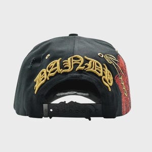 May include: Black baseball cap with gold embroidered text "BAND" across the back. The cap features a distressed design and a red and gold embroidered design on the side. The adjustable strap is black.