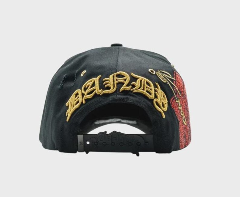 May include: Black baseball cap with gold embroidered text "BAND" across the back. The cap features a distressed design and a red and gold embroidered design on the side. The adjustable strap is black.