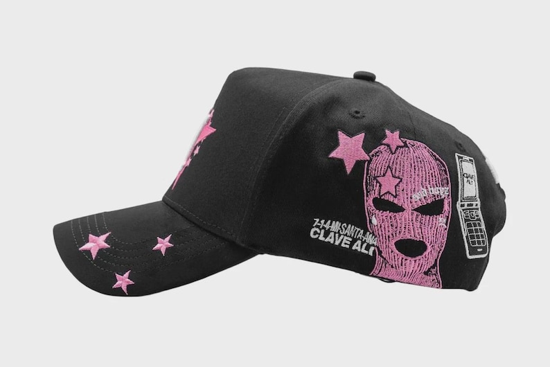 May include: Black baseball cap with pink star embroidery on the bill and side. The cap features a pink ski mask graphic and text that reads "7-14-MS SANTA-ANA CLAVE ALTO" and a flip phone graphic.
