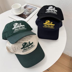 May include: A stack of baseball caps in various colors: black, navy, green, and beige. Each cap features the text "Aimé Dore Leon" and a graphic of a globe and a building. The caps are on a white surface.