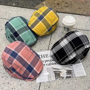 May include: A collection of plaid newsboy caps in various colors: green, yellow, pink, and black and white. The caps are arranged on a tiled surface with a newspaper clipping and a coffee cup.