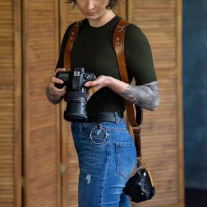 May include: A brown leather camera harness with a black camera and lens. The harness has shoulder straps and a chest strap. A small black camera case is attached to the harness. The person is wearing a dark green shirt and blue jeans.