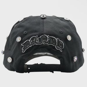 May include: Black baseball cap with the word "PARIS" embroidered in a gothic font. The cap is adorned with multiple clear, sparkling rhinestones. The adjustable strap is visible at the back.
